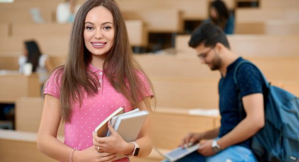 Pretty student keeping books in the modern spacy university classroom with wooden cascade desks. Standing on her groupmates background. Prepearing for exams. Smiling, feeling satisfied.
